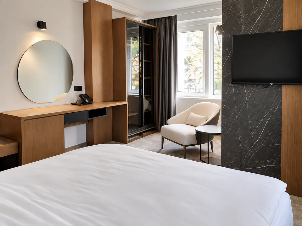 Apartment with a large bed, oak desk and round mirror, cream armchair by the window and a black marble TV wall