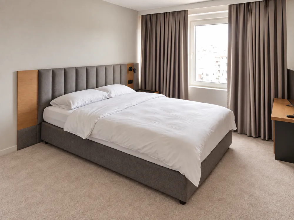 Double room with a large bed, grey padded headboard with oak panels, white linen and taupe curtains