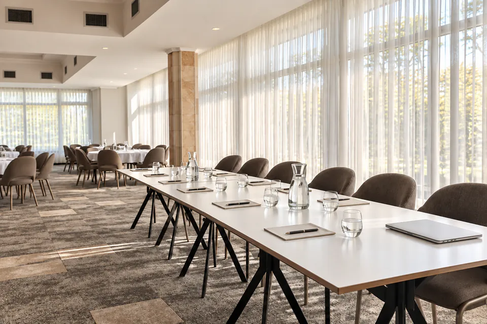 Seminar layout with a long white table, notepads, water carafes and upholstered chairs beside sheer-curtained windows